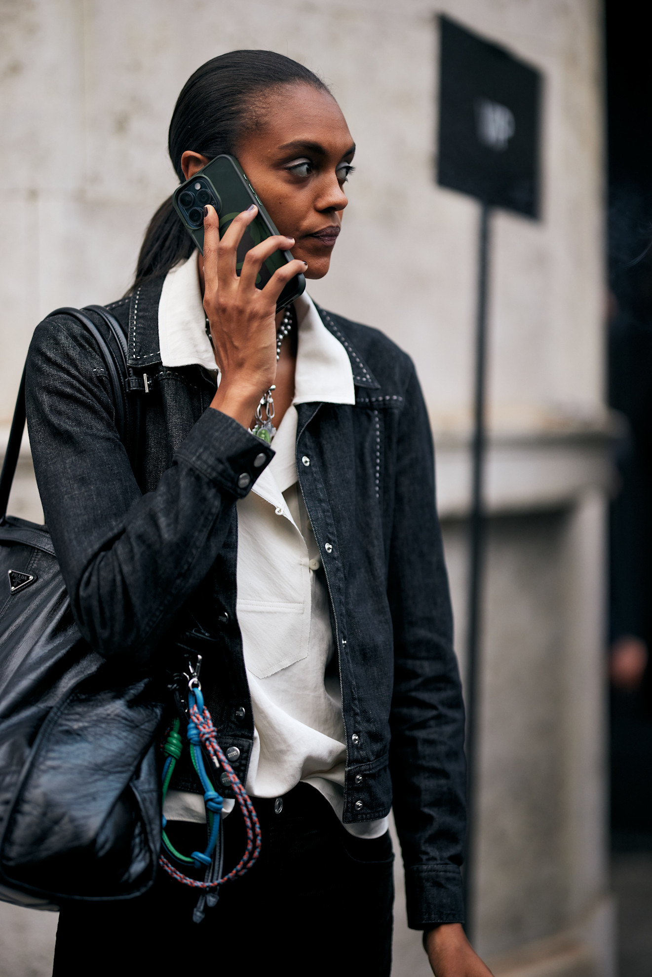 a woman with a slick-back ponytail and black nail polish wears a black denim jacket over a white blouse