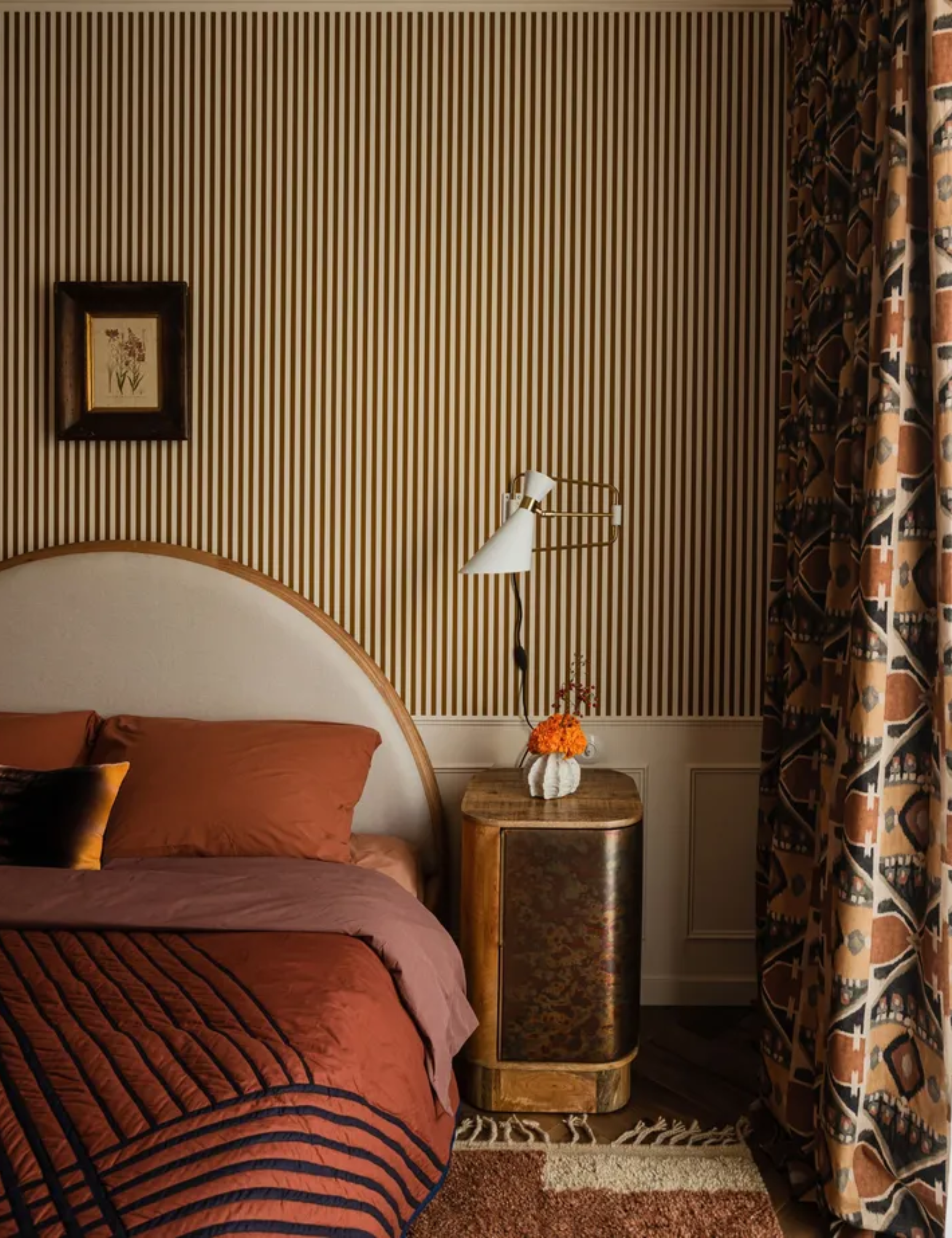 Art deco/ nouveau style bedroom with striped wallpaper, curved headboard and orange bedding, geometric curtains and burl wood and gold bedside table