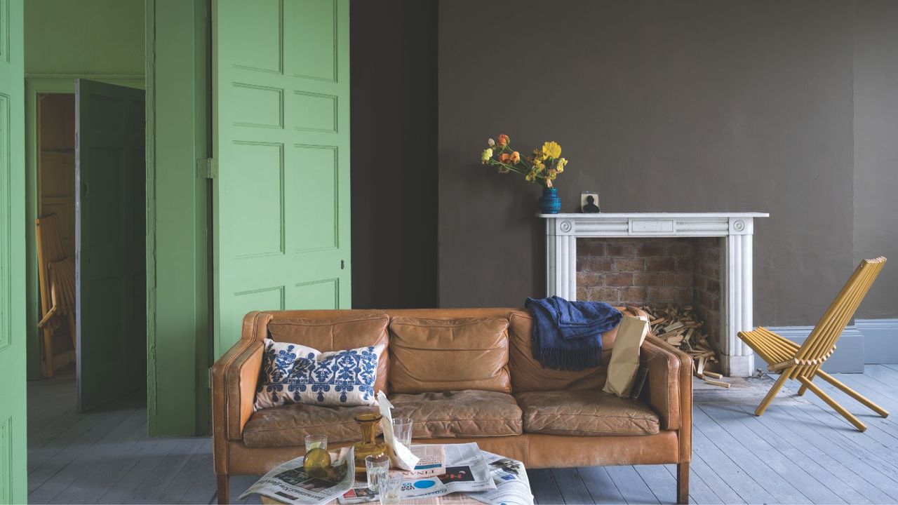 Living room with wooden floorboards, central wall painted dark brown around the large fireplace, a leather couch at the centre of the room, with a lime green painted door and hallway