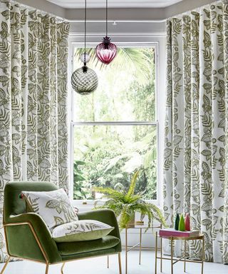 Green botanical floor length drapes in bay window, with colored glass pendant lights, and green velvet armchair with coord cushions.