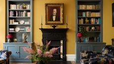 Living room with India Yellow walls by Farrow & Ball, a Victorian fireplace with a portrait above, built-in bookcases on either side painted grey-blue, and two velvet fabric armchairs.