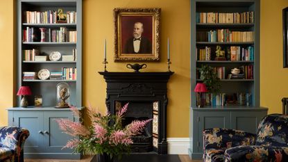 Living room with India Yellow walls by Farrow & Ball, a Victorian fireplace with a portrait above, built-in bookcases on either side painted grey-blue, and two velvet fabric armchairs.