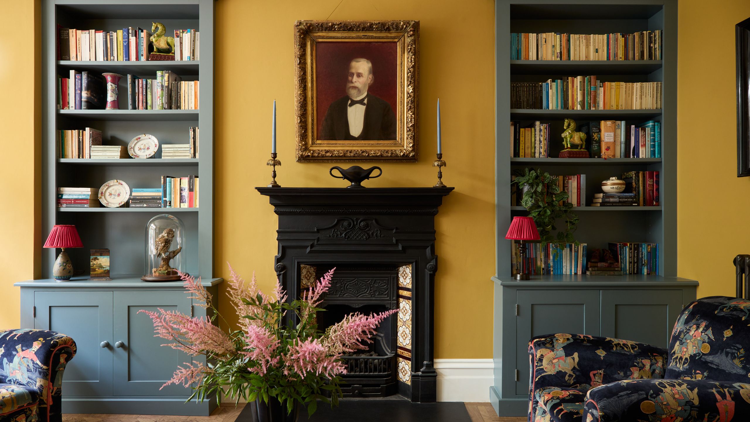 Living room with India Yellow walls by Farrow &amp; Ball, a Victorian fireplace with a portrait above, built-in bookcases on either side painted grey-blue, and two velvet fabric armchairs.