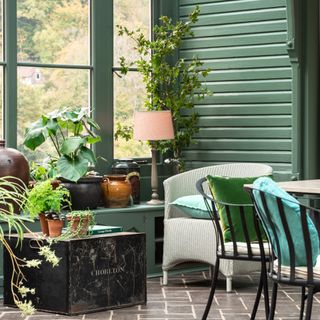 green painted conservatory interior with grey tiled floor, vintage chair, vintage chest, black metal chairs, green cushions plants, lamp