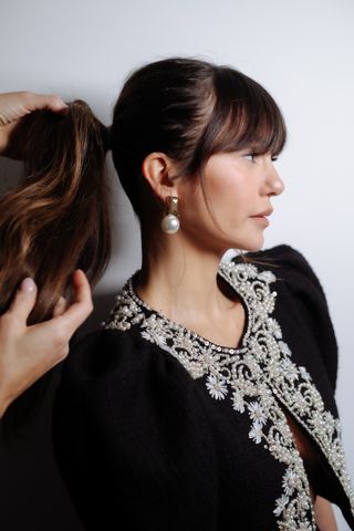 Nina Dobrev stands in front of a wall having her ponytail teased while wearing large drop earrings and a beaded shirt by Carolina Herrera