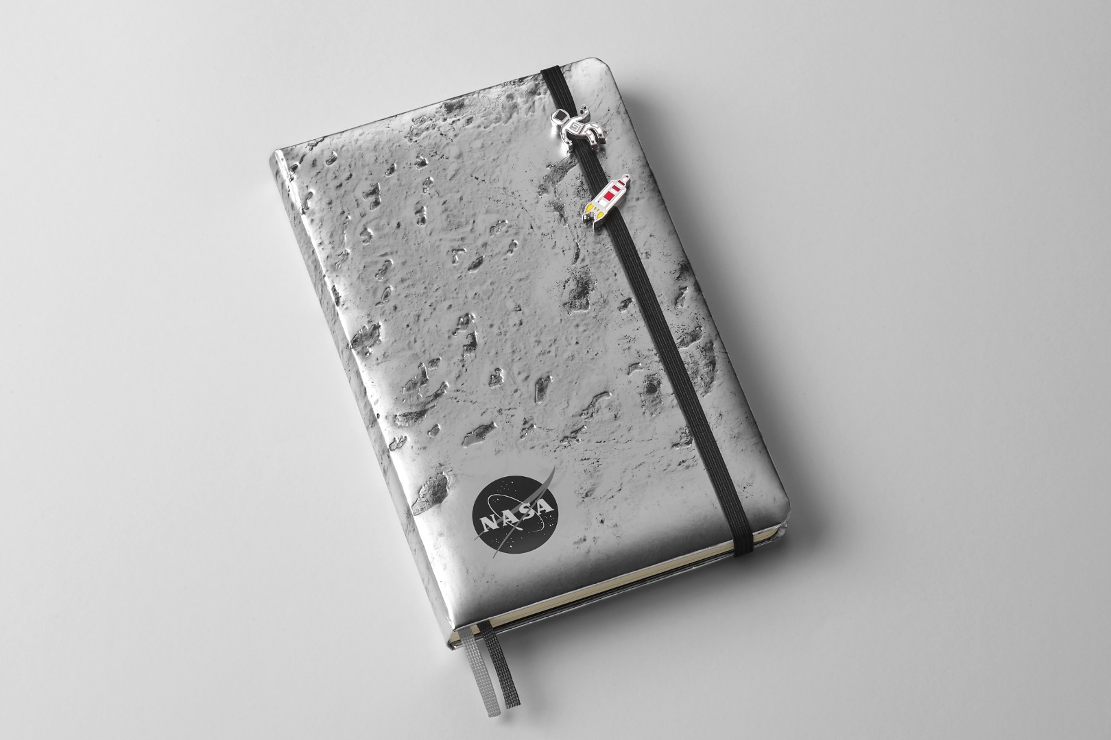 Two small pins attached to the black elastic closure of the NASA-inspired Limited Edition Notebook from Moleskine (silver moon surface). The top pin is a person in a white astronaut suit with red trim. The bottom pin is a small grey, white, and red simplistic rocket blasting off.