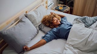 A woman with afro hair wearing blue shirt waking up in three quarter bed with grey sheets, stretching our arms