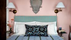 Blush pink painted bedroom with vintage-looking patterned blue bedding, striped green and cream headboard with paired brass wall lamps and an ornate wall mirror