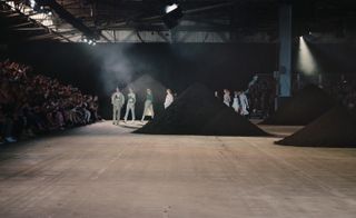 Female models walking on a wooden stage with large mounds of dark sand on it and a crowd sitting on a stand next to it.