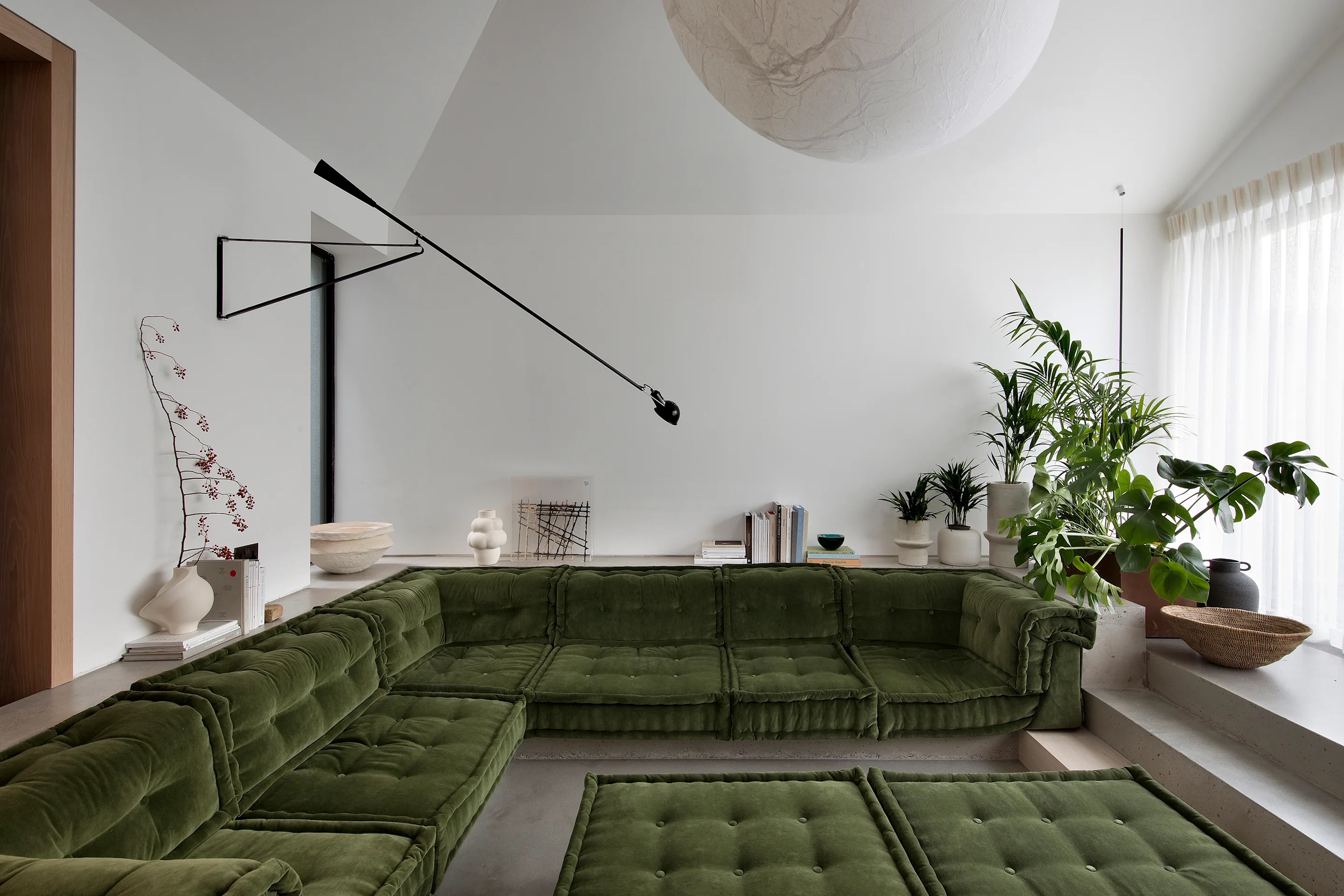 A sunken seating area in dark green with polished concrete floors, white walls, a wall-mounted light, vases styled around the room, and a corner filled with houseplants