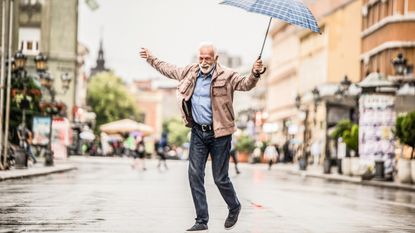 An older man happily dances in the rain under an umbrella.