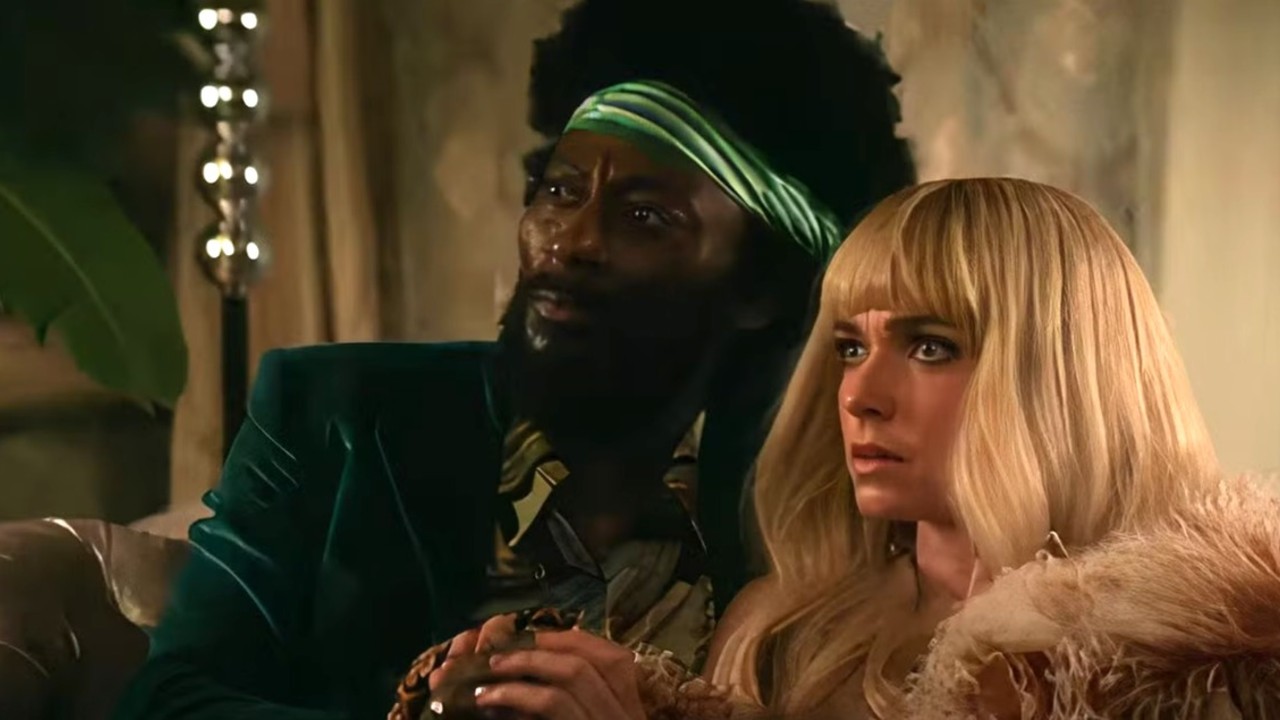 a man and a woman in 1970s-era wigs sit on a couch