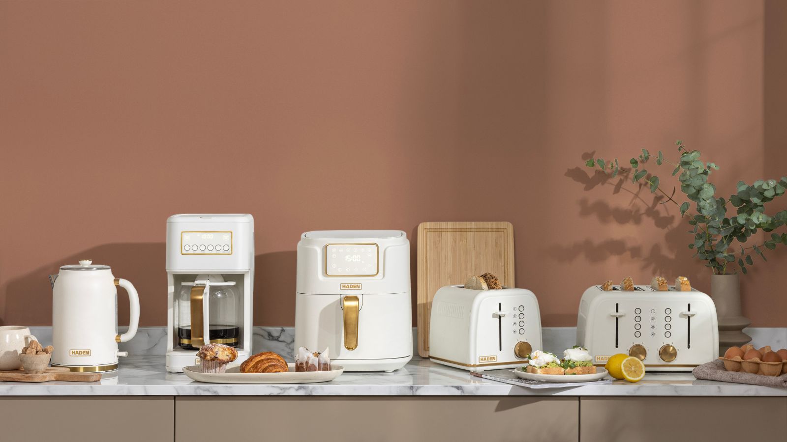 Mocha-colored kitchen with white marble counters and a set of white appliances (kettle, coffee maker, air fryer, and two toasters) all with brass accents