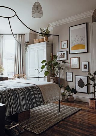 A beige bedroom with gallery wall, potted houseplants, white closet and four-poster bed with grey and white rug on wooden flooring