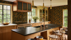 a kitchen with dark floral wallpaper, black stone countertops, a custom copper range hood, and light wood cabinetry