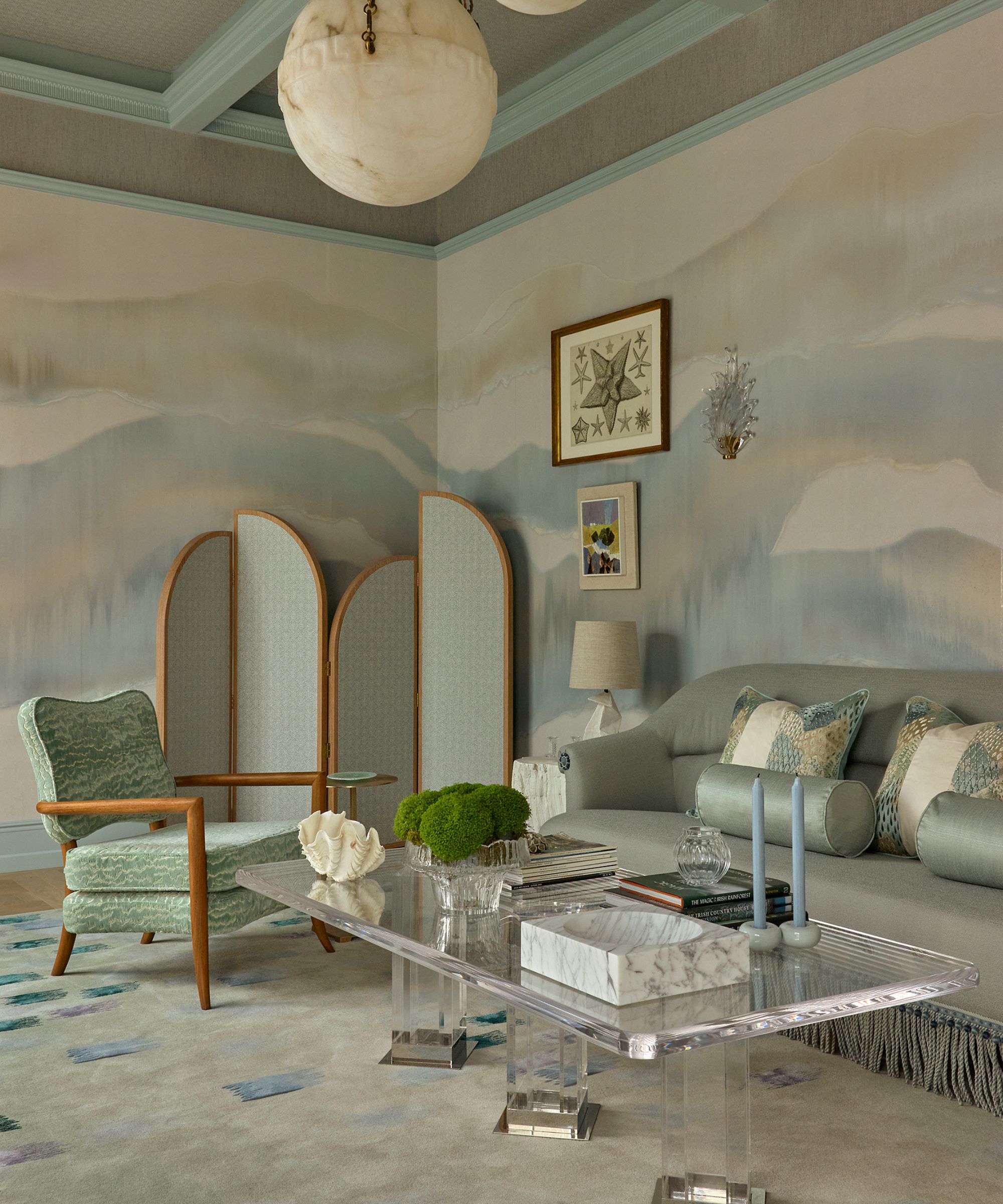 A seafoam-painted morning room with abstract handpainted walls, a blue-painted ceiling, a glass pendant light, a blue fabric screen, a silk blue sofa with fringe, and glass coffee table