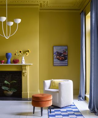 how to design a room that feels like you, yellow living room with matching ceiling, white painted floorboards, white boucle armchair, blue and white rug, orange footstool, blue drapes, vests on mantel, modern white pendant, artwork