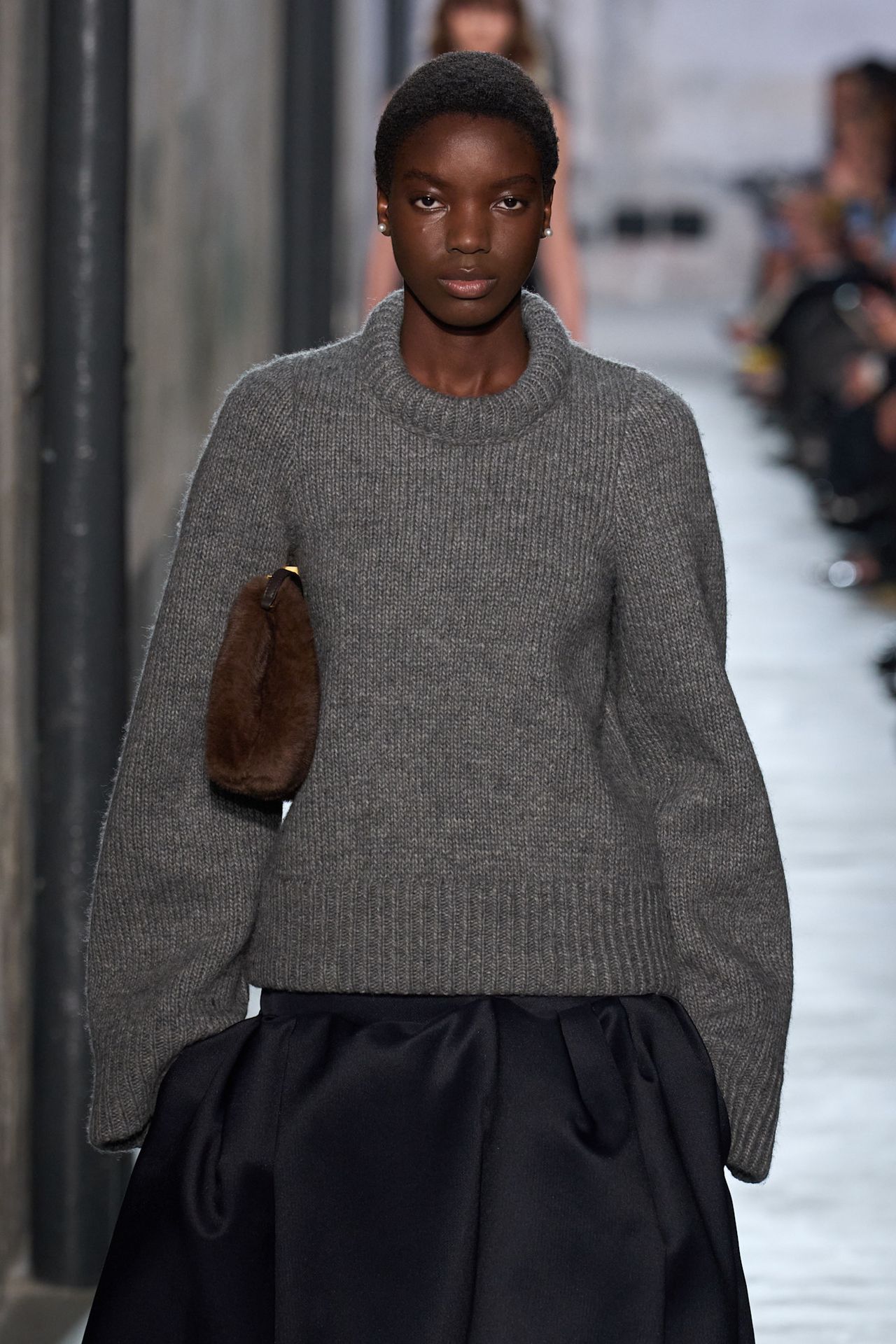 Fur accessory trend is shown in N&ordm;21's fall-winter 2025 collection in a close photo of a model walking down the runway wearing a gray high-neck sweater with a black drop waist voluminous skirt and a dark brown fur clutch bag