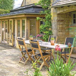 exterior image of wooden orangery with slate roof attached to a stone property with patio area and wooden table and chairs