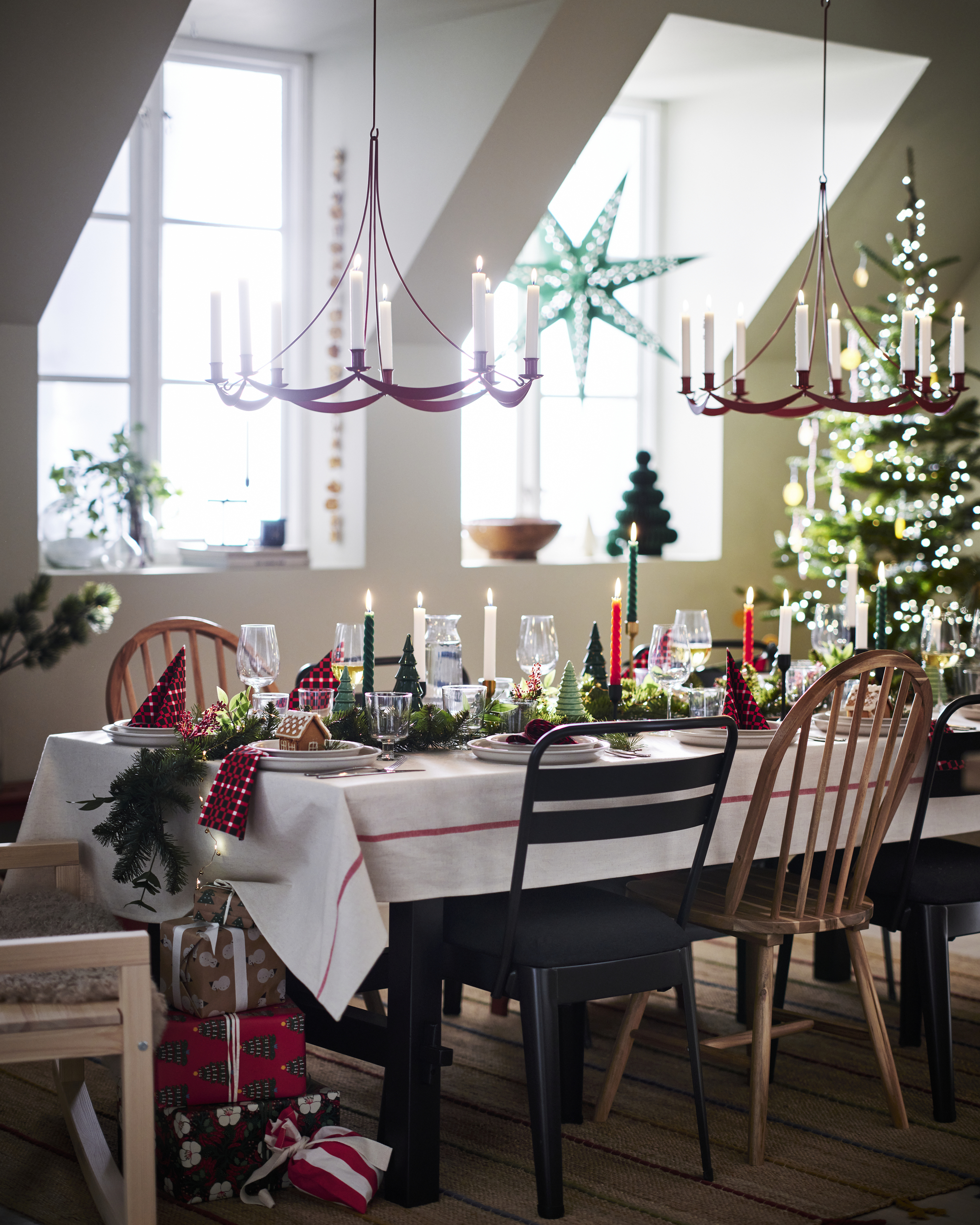 Image of a dining room decorated for Christmas. There is a tree in the corner, two windows on the back wall, and a table decorated with holiday decor and two red candle chandeliers hanging over it.