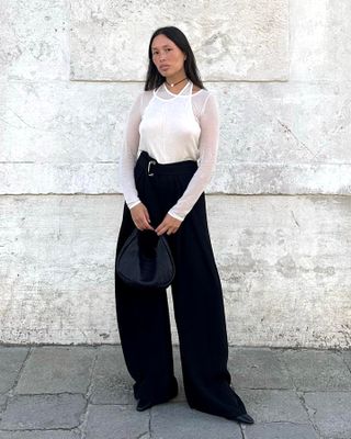 French influencer Anne-Victoire Lefevre poses wearing a sheer white layered top, belted wide-leg black trousers, and a mini black bag.