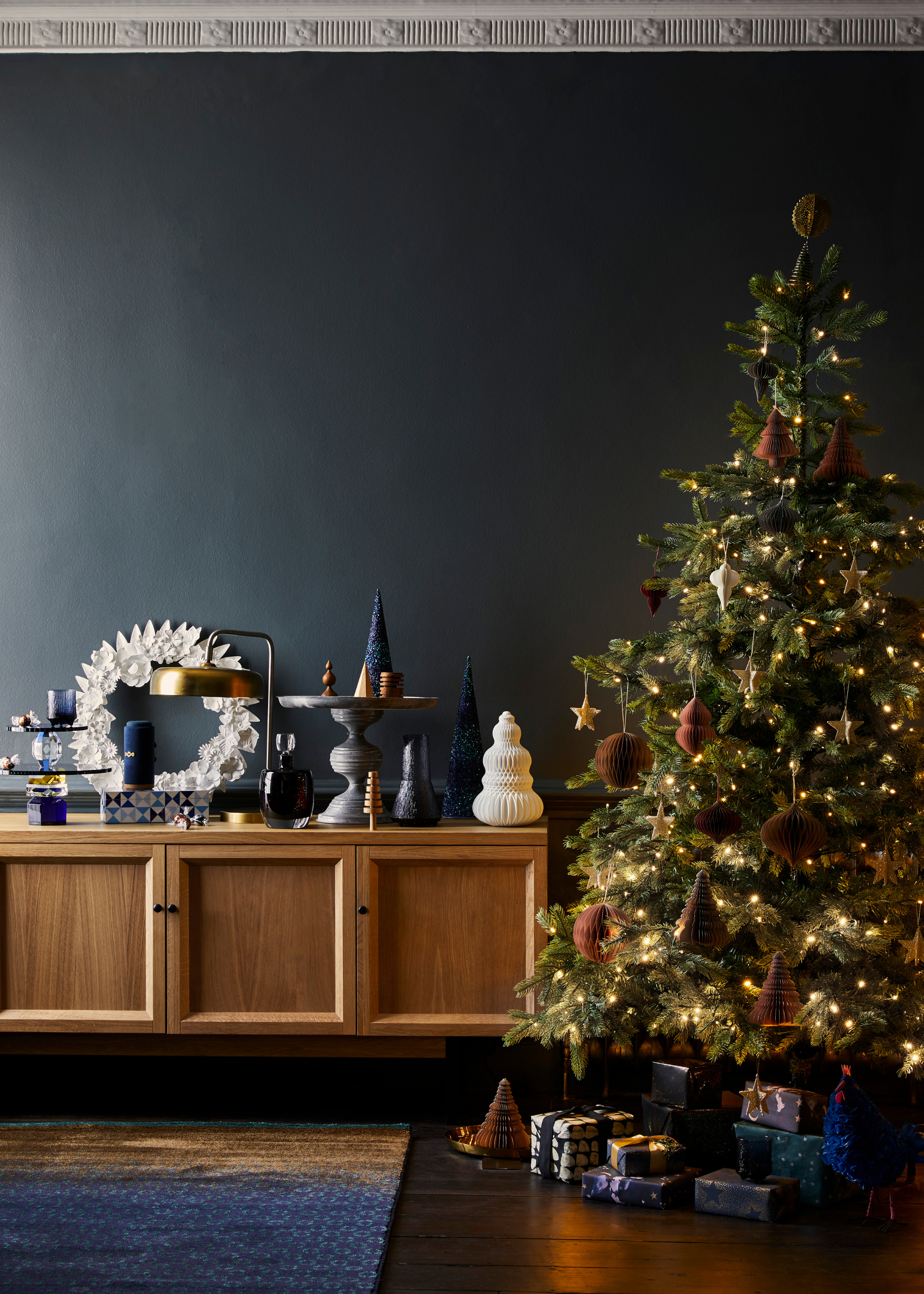 A christmas tree with gold and brown decorations and a natural wood console table contrast a dark blue-painted wall and rug and the decorations on top of the console table are dark blue, gold and white with some decorations elevated on a stand