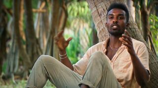 Kyle sits next to a tree and raises his hands, talking about his strategy.