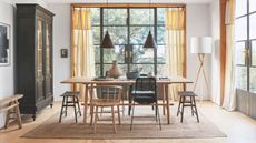 Bright and airy dining area with floor-to-ceiling windows, a wooden dining table, mismatched chairs and a large jute rug. To the left is a large black cabinet, and to the right is a tall wooden lamp with white lampshade.