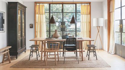Bright and airy dining area with floor-to-ceiling windows, a wooden dining table, mismatched chairs and a large jute rug. To the left is a large black cabinet, and to the right is a tall wooden lamp with white lampshade.