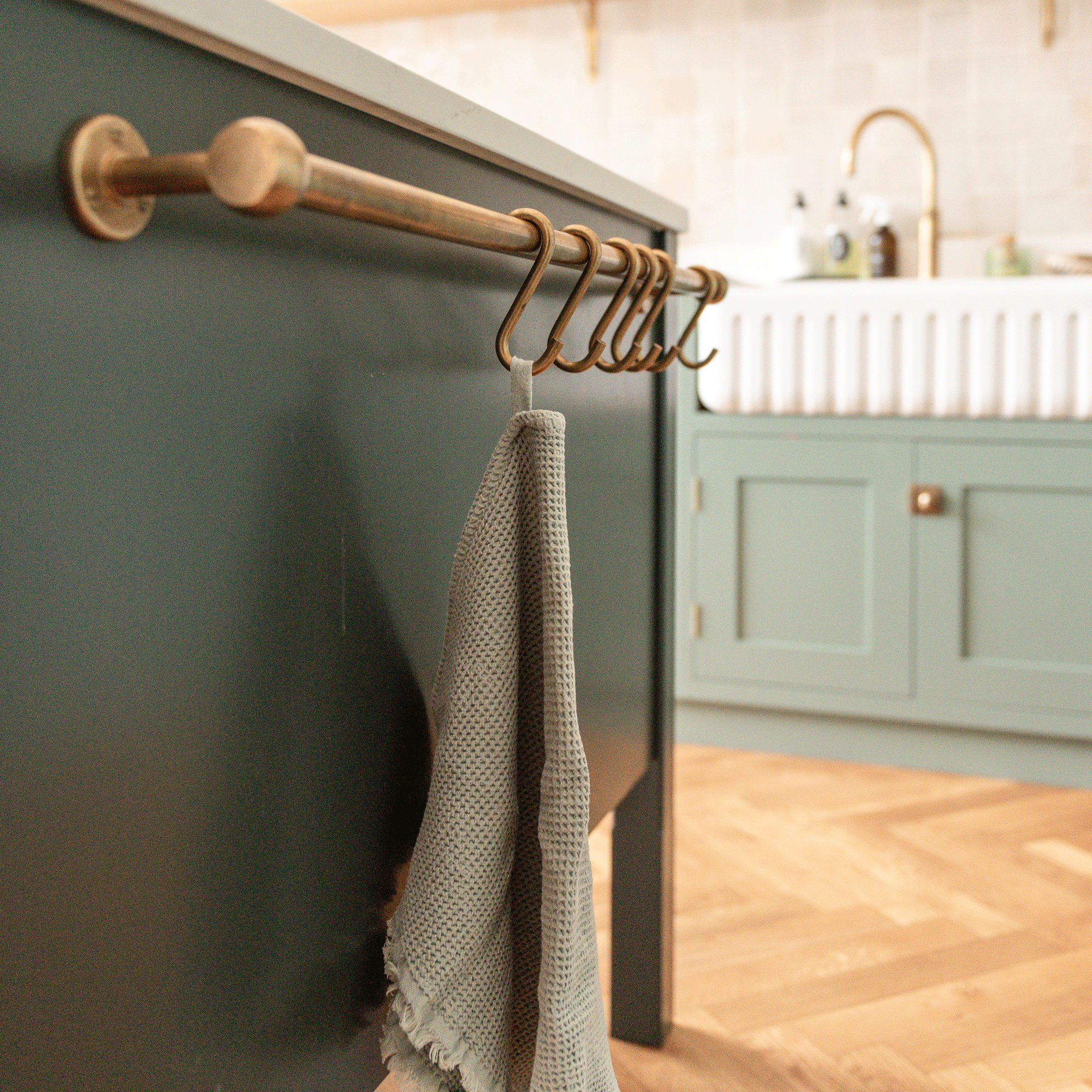 close up of a kitchen island in a sage green kitchen with a brass rail and s-hooks for tea towels