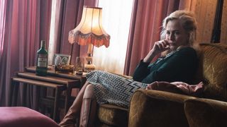 Gillian Anderson as Gina sitting in a brown arm chair with a bottle of drink beside her looking upset.