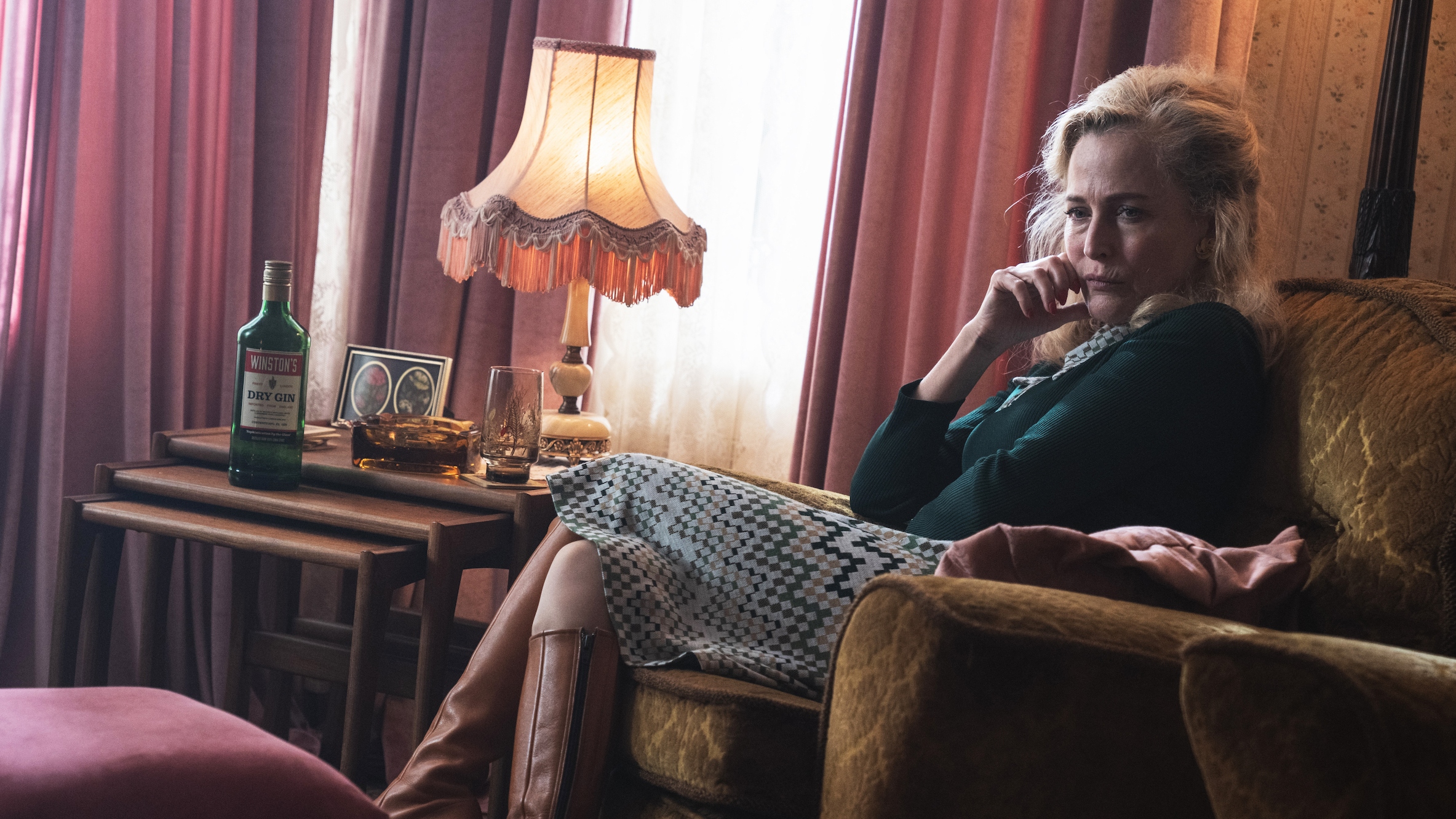 Gillian Anderson as Gina sitting in a brown arm chair with a bottle of drink beside her looking upset. 