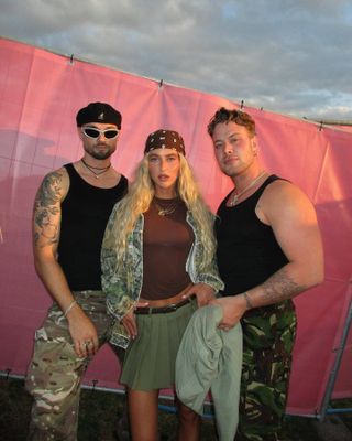 Emili stands with two men wearing a green pleated mini skirt, a black belt, brown top, camo jacket, brown bandana, and gold necklace.