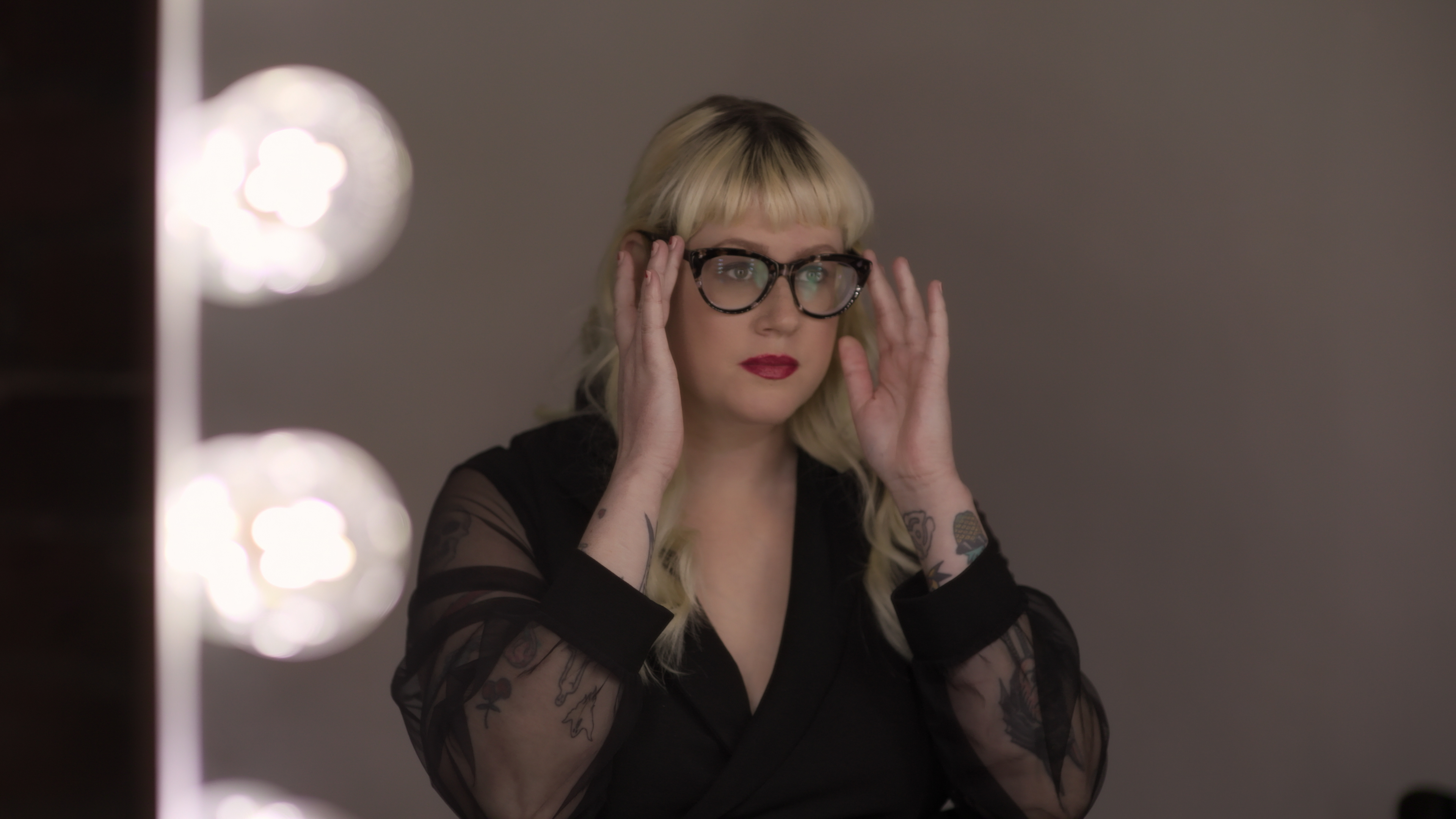 A blond woman wearing all black (Shandi Sullivan) looks into a vanity mirror while adjusting her glasses, in a still from the Netflix doc 'Reality Check: Inside America's Next Top Model.'