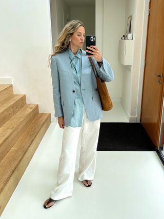 Anouk Yve taking a mirror selfie wearing a blue shirt and summer blazer, white wide-leg trousers, flip-flops, and carrying a brown suede tote.