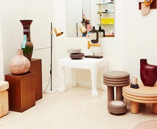 A room with a range of furniture and tableware items, including two brown side tables with vases on top. A standing lamp. A white table with plates on. A large mirror on the wall. Right: two different style stools, one tall and skinny, one low and round.