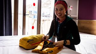 A woman sits in a hotel room, packing clothes in two yellow Eagle Creek Pack-It cubes.