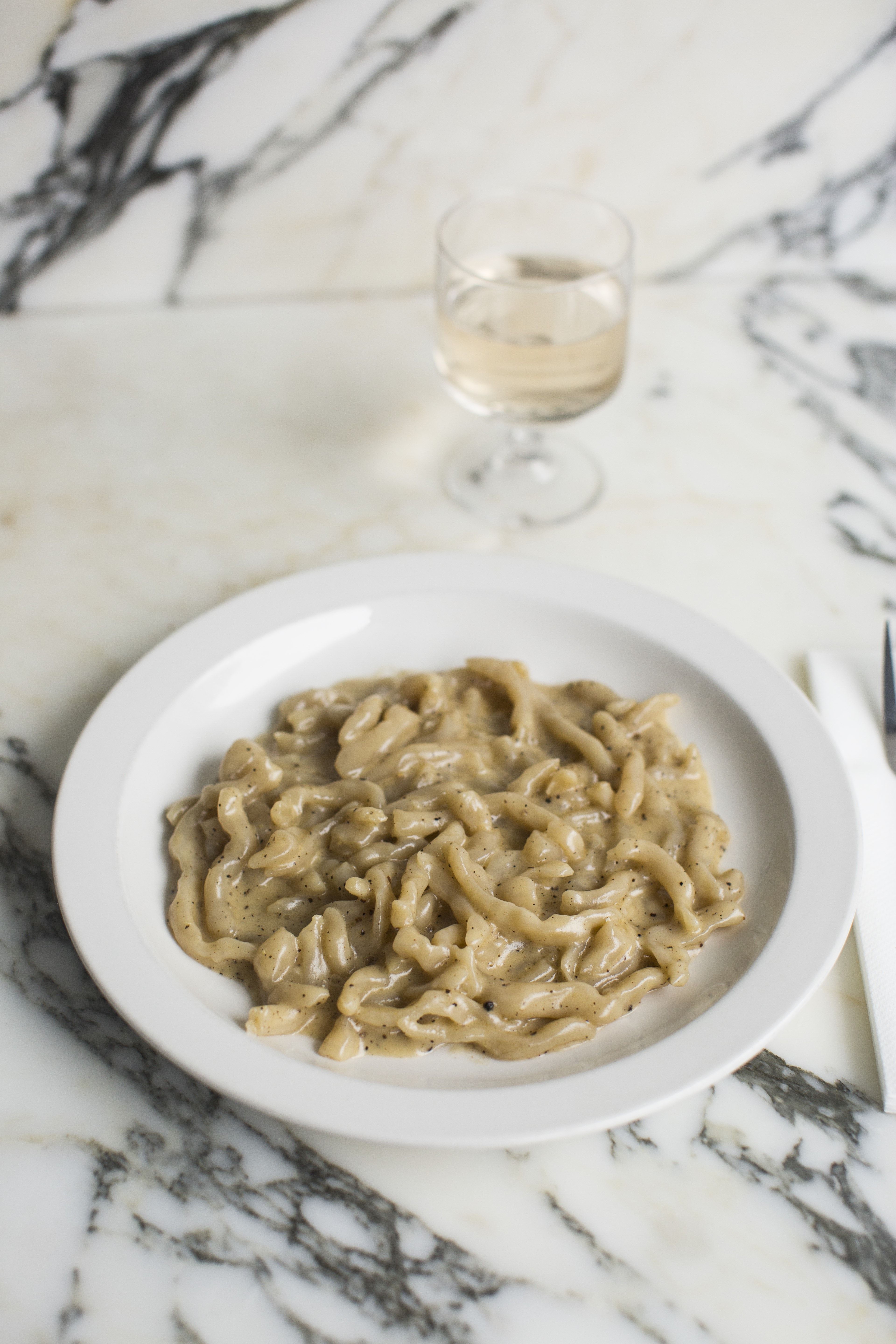A plate of creamy mushroom pasta in captured against a marble countertop next to a glass of white wine.