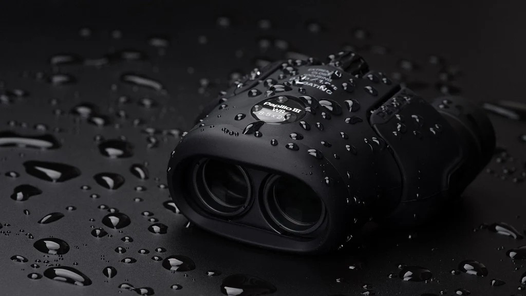 A pair of black Pentax Papilio III 6.5x21 WR binoculars is shown resting on a wet black surface, demonstrating its water-resistant properties with water droplets covering the body.