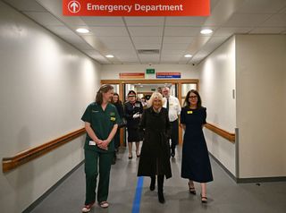 Queen Camilla walking down a hospital hallway with staffers with a red sign reading Emergency Department above