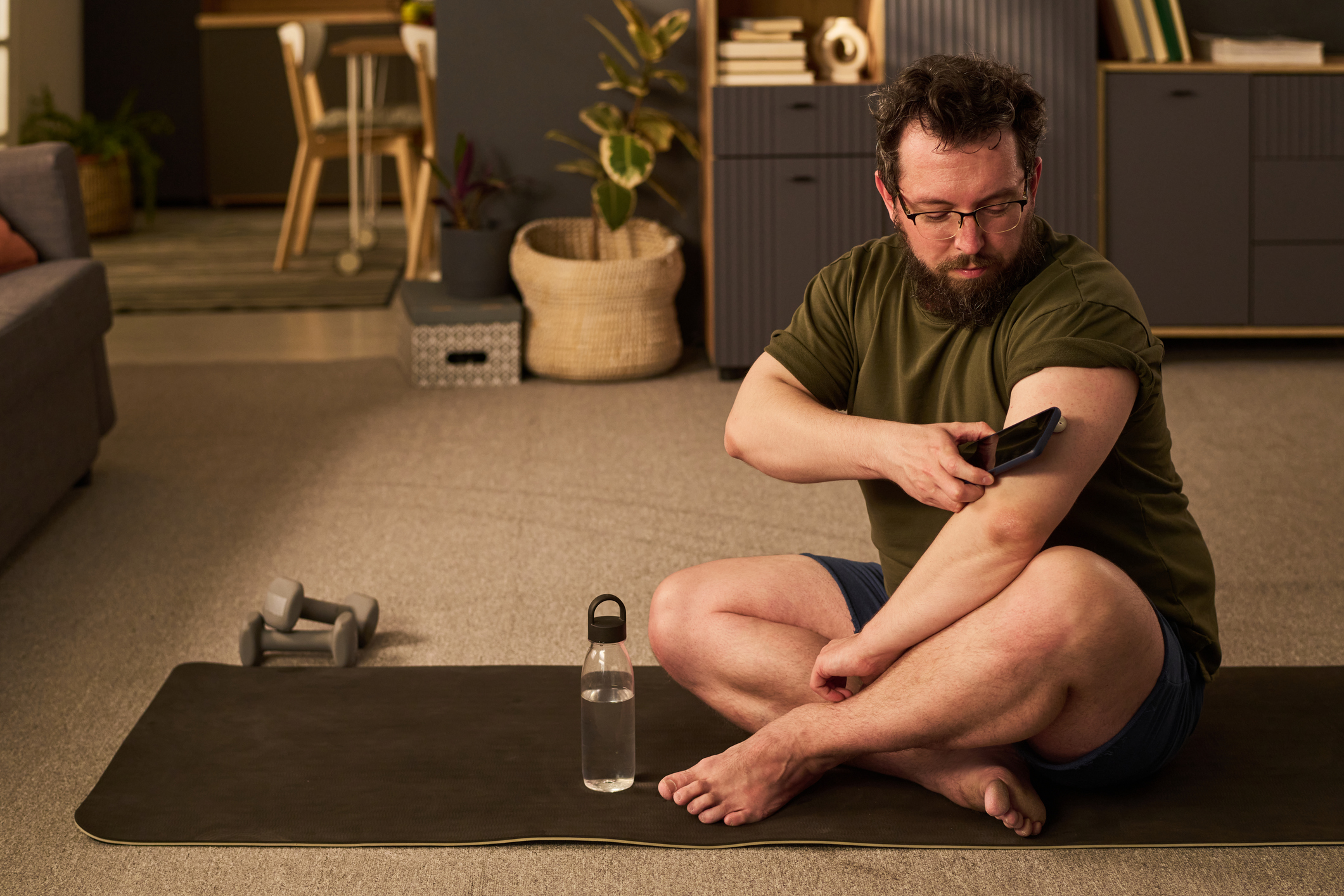 A man in shorts and T-shirt sits on a yoga matter with his legs crossed, he holds a smartphone next to a glucose monitor on his arm