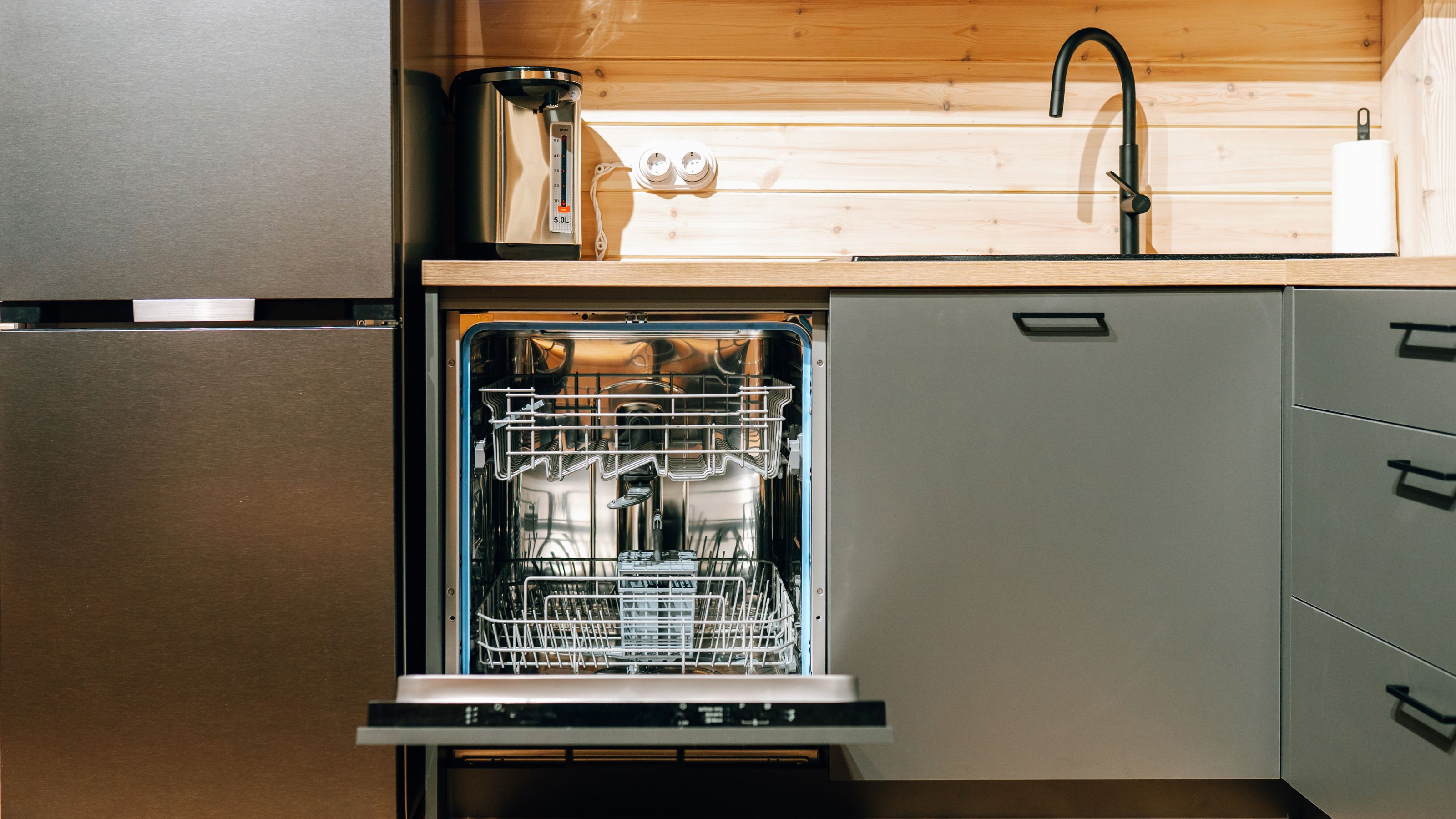 Open integrated dishwasher next to a fridge freezer, in a kitchen with wood panelling, a wood-look worktop, and sage green cabinet doors and black handles