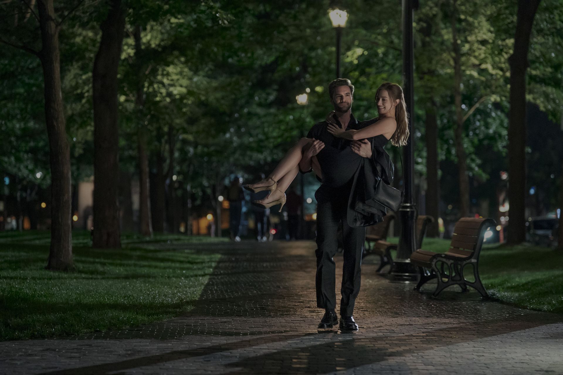 tom bateman as adam carrying lili reinhart as olive down a dark path in a park on a date in the love hypothesis