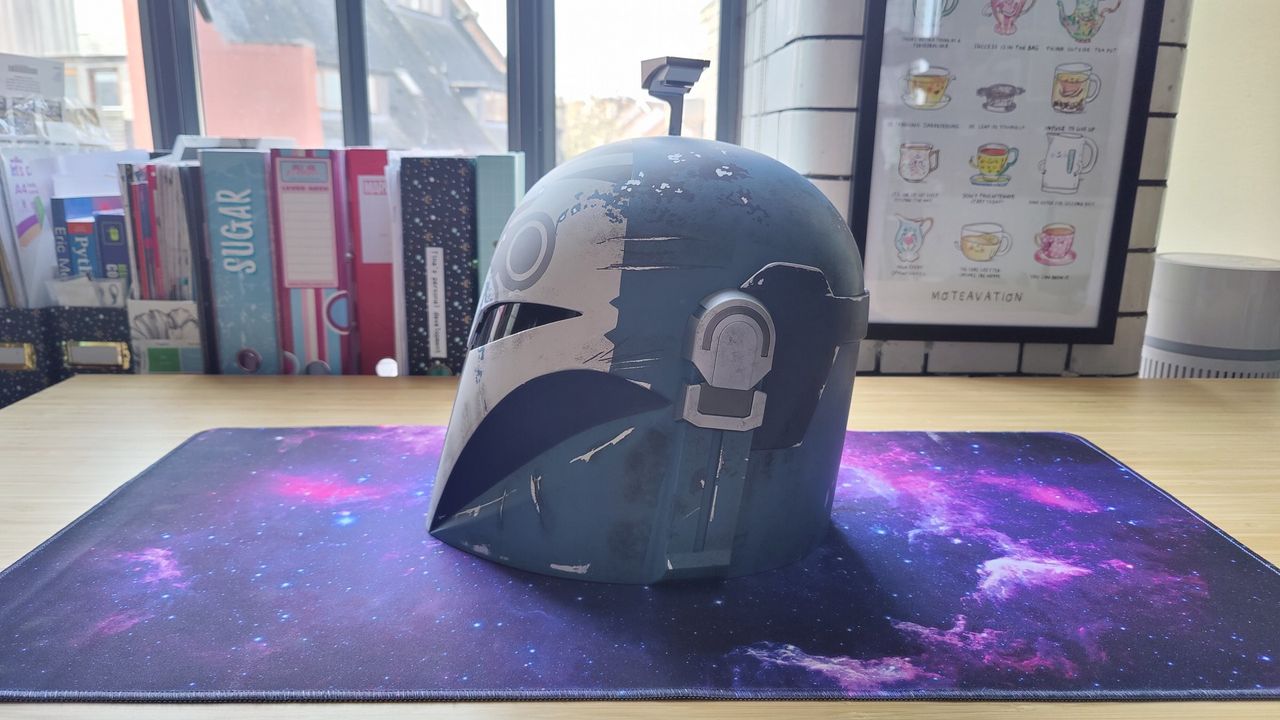 The Star Wars: Black Series Bo-Katan Helmet with the heads-up display up. There is a gray, black, and white owl-like pattern on the front of the light blue helmet and a black T-shaped visor. This photo shows the left-side of the helmet.