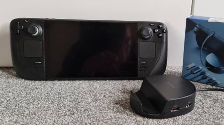 Syntech dock next to Steam Deck on grey carpet.