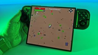 Hand holding a wide foldable smartphone displaying active Brotato gameplay, clamped horizontally inside a black Razer Prio controller under green light.
