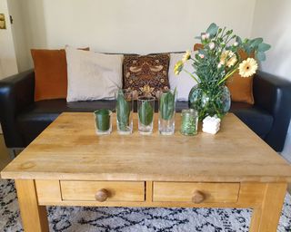 Joanna Gaines' budget centerpiece hack demonstrated on a coffee table - showing leaves places inside of glasses, with a floral arrangement on the side, and a sofa beyond