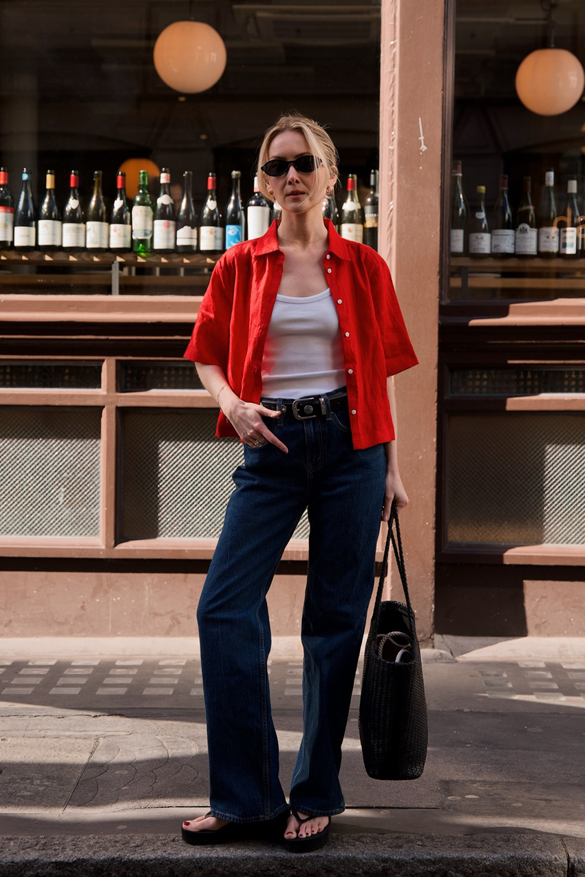 A woman wearing red short sleeve button-down, white tank top, dark-wash denim jeans, and black wedge sandals.