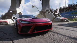 A red racecar in Forza Horizon 6 idling on a racetrack in front of the massive metal feet of an otherwise invisible mecha.