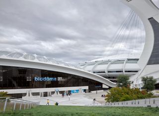 Montreal Biodome's curved white exterior, glass fronted entrance with blue biodome sign, surrounding grass verge and trees on a cloudy day